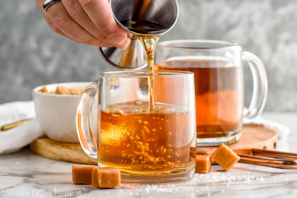 man's hand pouring dark rum into a mug of salted caramel buttered rum ingredients with mug of salted caramel buttered rum and bowl of brown sugar sitting in background and caramel candies sitting beside.