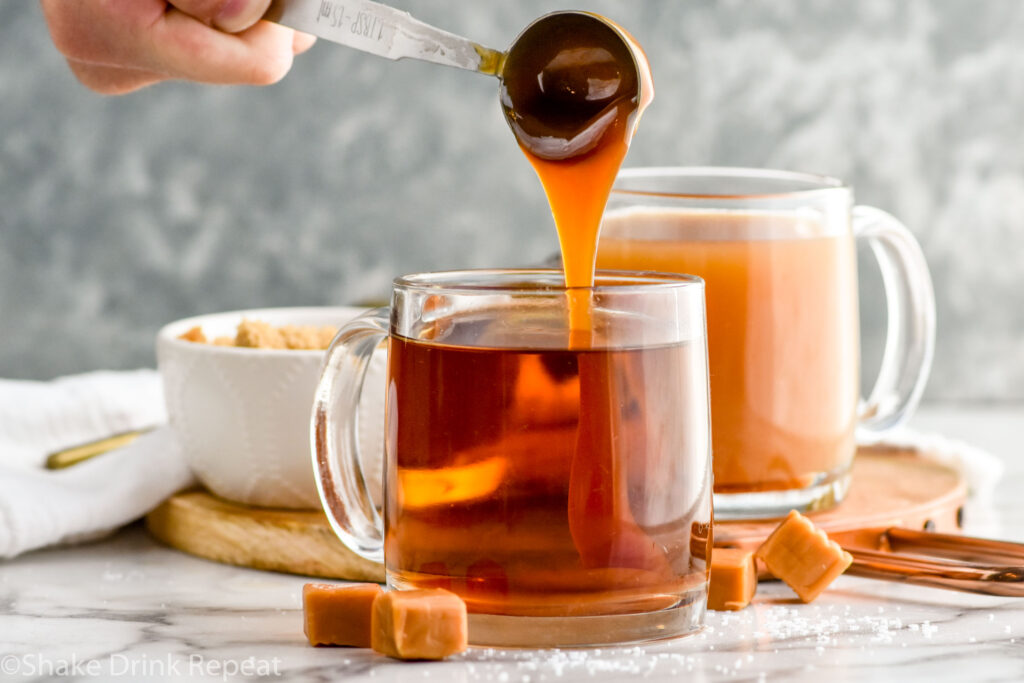 man's hand pouring tablespoon of caramel sauce into a mug of salted caramel buttered rum cocktail with mug of salted caramel buttered rum and bowl of brown sugar sitting in background and caramel candies sitting in background