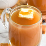 Mug of salted caramel buttered rum with bowl of brown sugar sitting in background and caramel candies sitting beside.