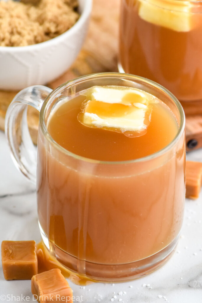 Mug of salted caramel buttered rum with bowl of brown sugar sitting in background and caramel candies sitting beside.