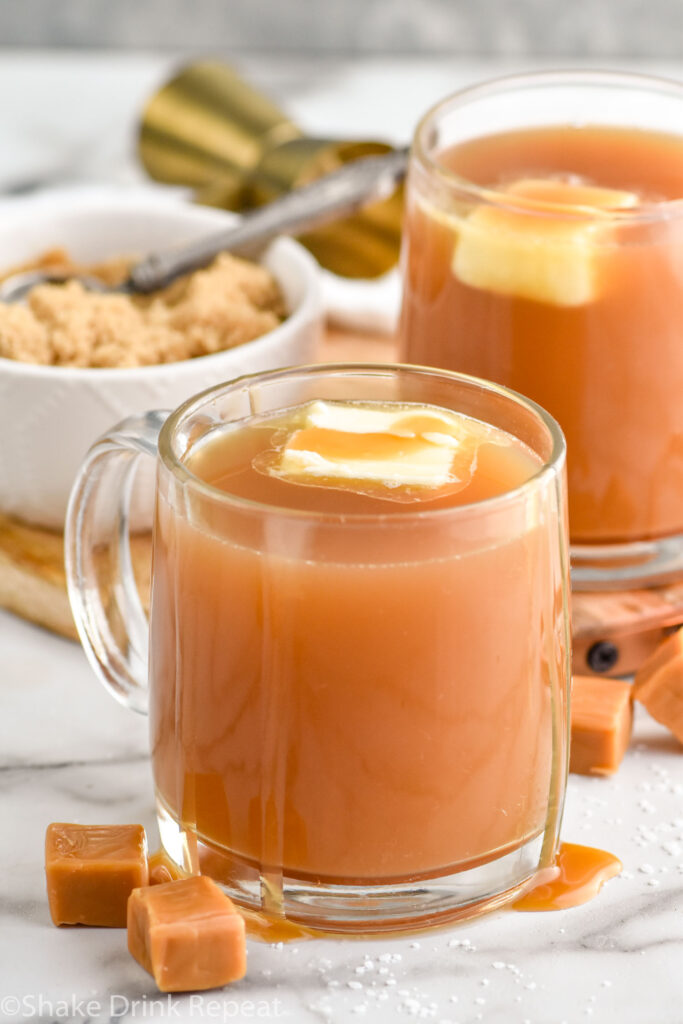 Two mugs of salted caramel buttered rum with bowl of brown sugar sitting in background and caramel candies sitting beside.