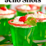 Pinterest graphic of Drunken Elf Jello Shots with a front facing photo of a jello shot with whipped cream, a red hot, and mint with more shots behind it on a golden stand. Says, 'So Easy, Drunken Elf Jello Shots, shakedrinkrepeat.com'