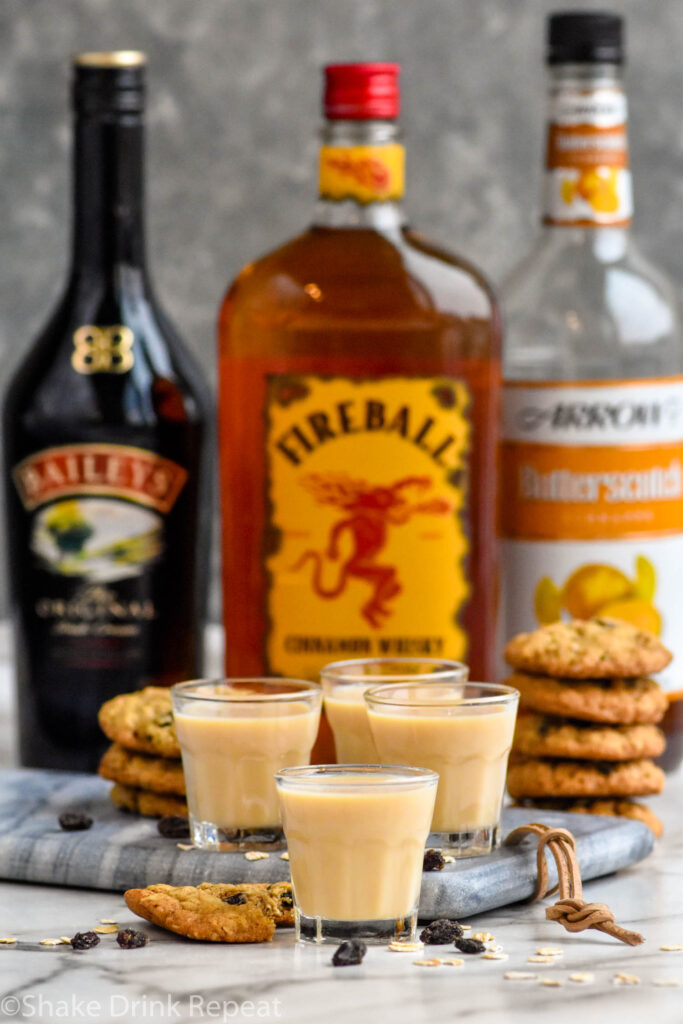 Front facing photo of Oatmeal Cookie Shot with three shots that have Baileys, Fireball, and Butterscotch Schnapps bottles behind it and stacks of oatmeal cookies.