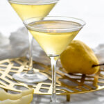 Front facing photo of Pear Martini with two glass of Pear Martini in martini glasses on a gold stand and a pear with pear slices around it.