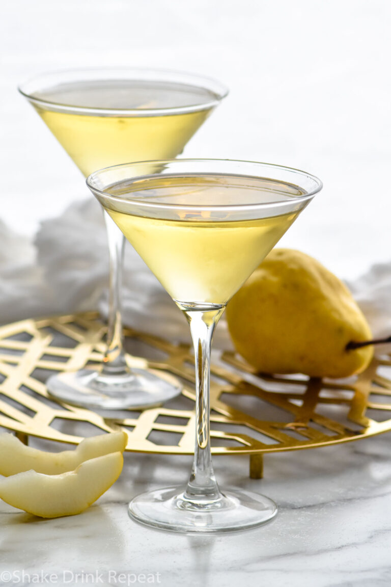 Front facing photo of Pear Martini with two glass of Pear Martini in martini glasses on a gold stand and a pear with pear slices around it.