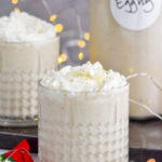 Front facing photo of Spiked Eggnog with a second glass of eggnog to the back left and a bottle of eggnog to the back right with sparkling lights behind it.
