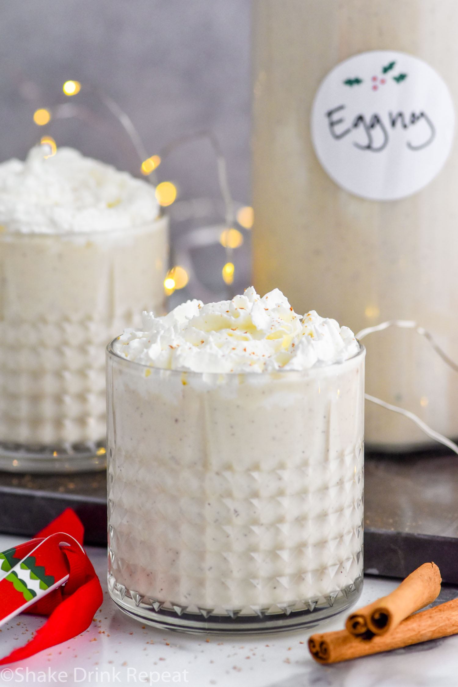 Front facing photo of Spiked Eggnog with a second glass of eggnog to the back left and a bottle of eggnog to the back right with sparkling lights behind it.