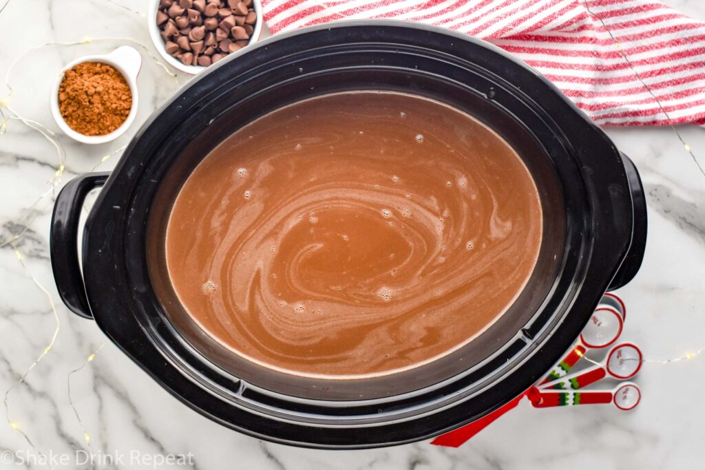 overhead of crockpot hot chocolate. Measuring spoons, cocoa powder, and chocolate chips sitting beside.