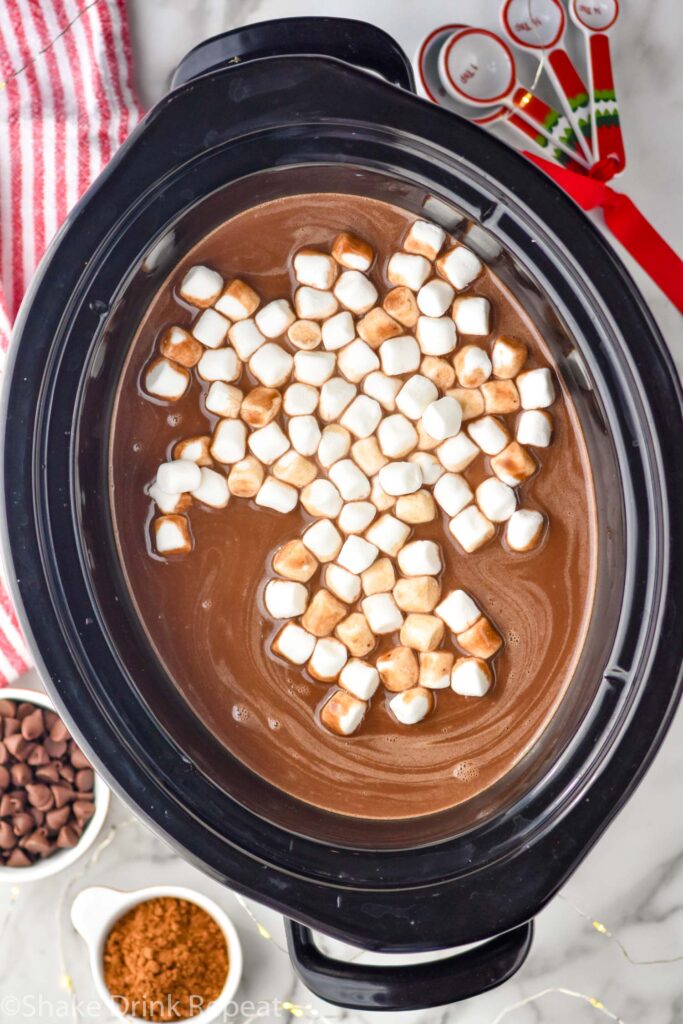 overhead of crockpot hot chocolate topped with marshmallows. Measuring spoons, cocoa powder, and chocolate chips sitting beside.