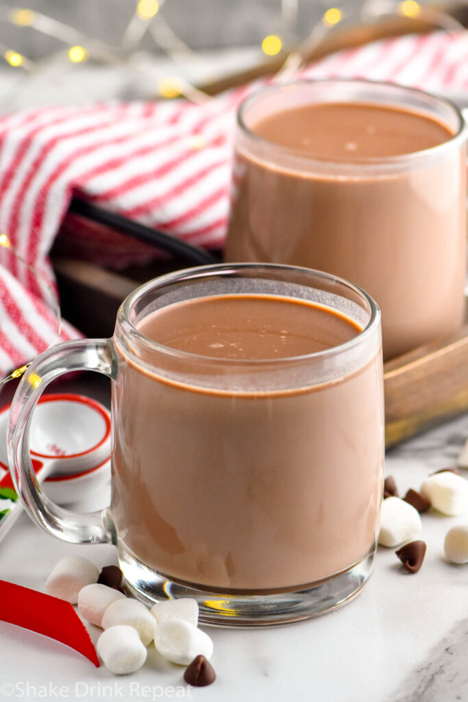 two mugs of crockpot hot chocolate. Marshmallows and chocolate chips in front of mugs