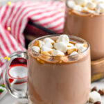two mugs of crockpot hot chocolate topped with marshmallows. Marshmallows and chocolate chips in front of mugs