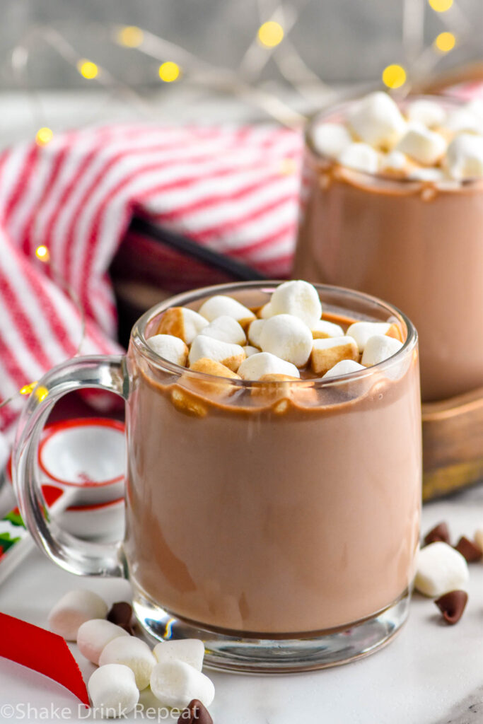 two mugs of crockpot hot chocolate topped with marshmallows. Marshmallows and chocolate chips in front of mugs