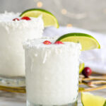 Front facing photo of White Christmas Margarita with cranberries and a lime wedge garnish and extras around the glass with another margarita glass in the background.