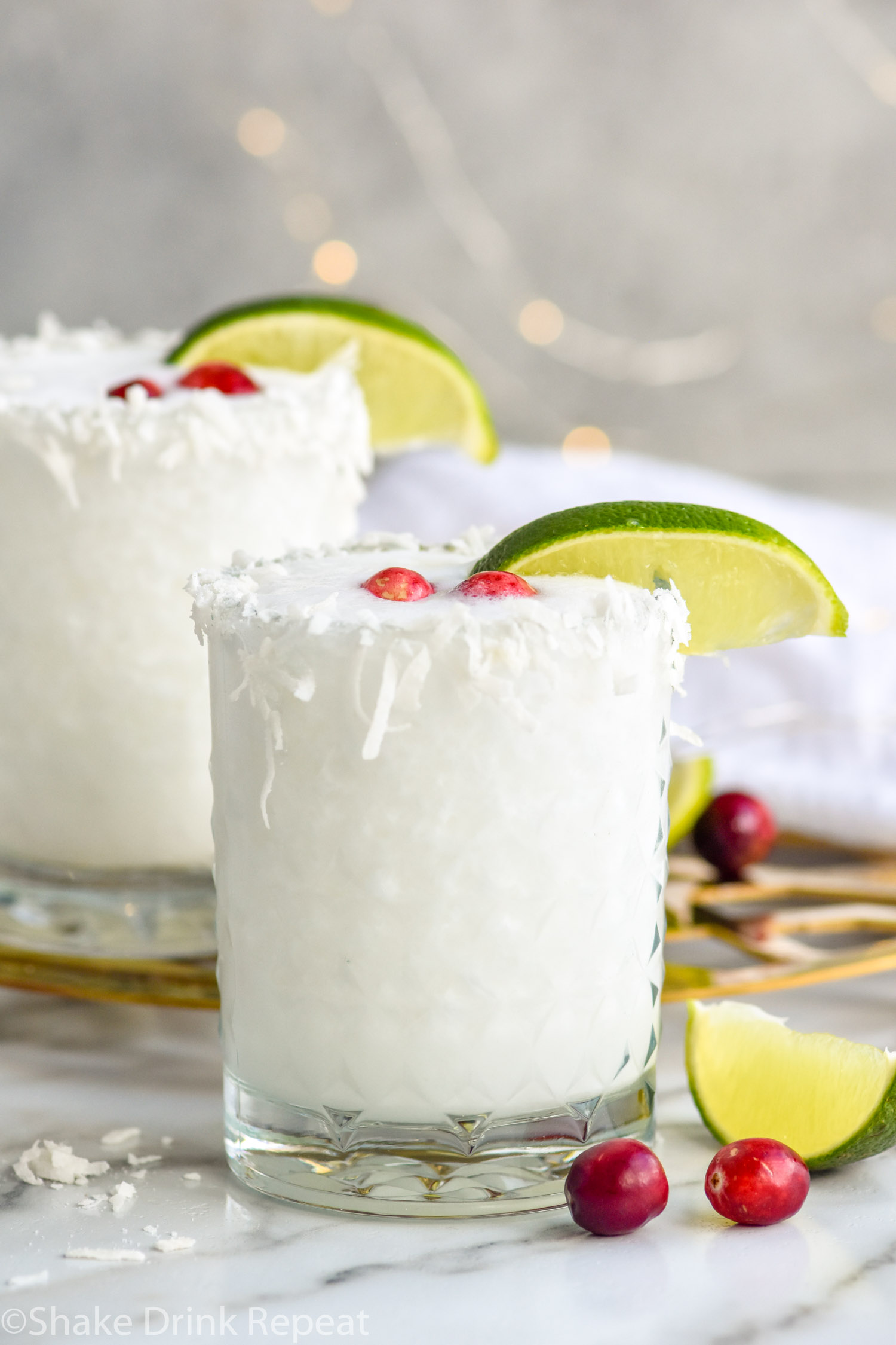 Front facing photo of White Christmas Margarita with cranberries and a lime wedge garnish and extras around the glass with another margarita glass in the background.
