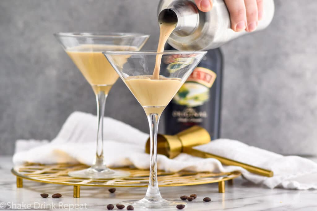 Close up photo of person's hand pouring Baileys Espresso Martini from a cocktail shaker into a martini glass. Second Baileys Espresso Martini and bottle of Baileys on also on counter.