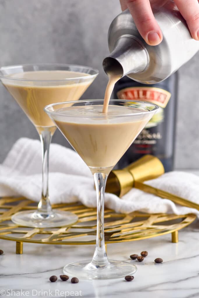Photo of person's hand pouring a Baileys Espresso Martini into a glass. Bottle of Baileys and a second Baileys Espresso Martini sit on counter behind cocktail.