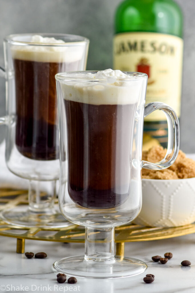 Close up photo of two mugs of Irish Coffee topped with whipped cream. Bowl of brown sugar and bottle of Jameson Irish Whiskey on counter behind mugs.