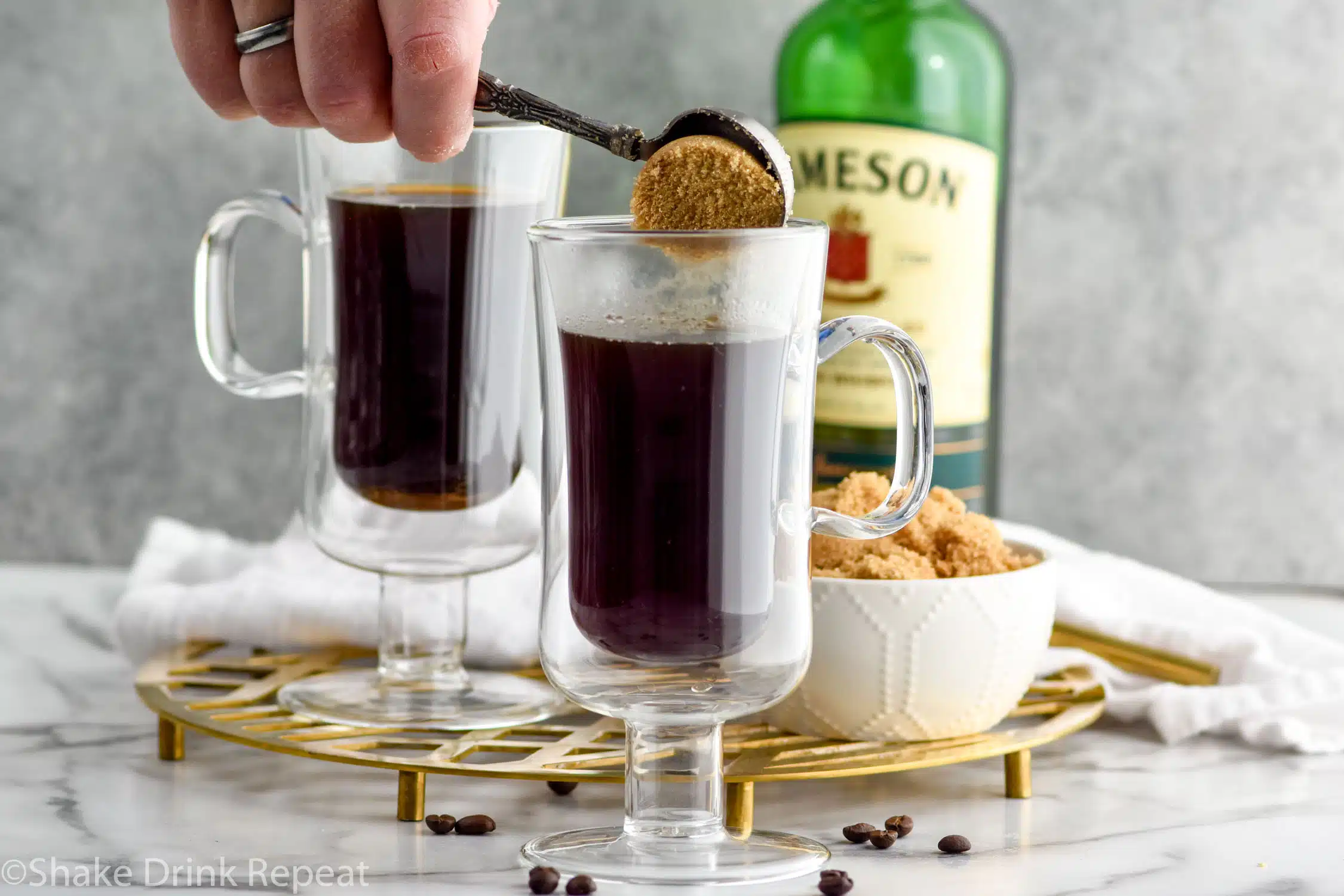 Photo of person's hand spooning brown sugar into mug of coffee for Irish Coffee recipe. Bottle of Jameson Irish Whiskey and a bowl of brown sugar sit on counter behind mugs.