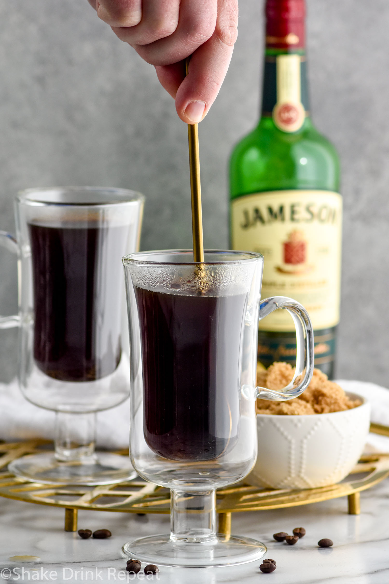 Photo of two mugs of coffee for Irish Coffee recipe. Person's hand shown stirring one mug. Bowl of brown sugar and a bottle of Jameson Irish Whiskey in the background.