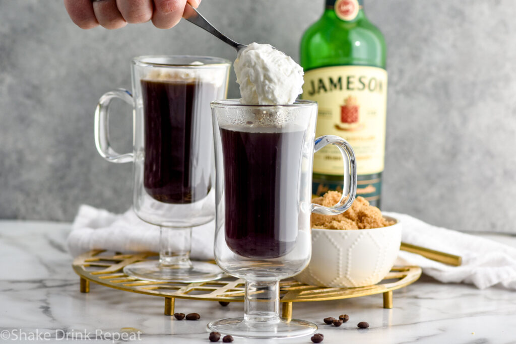 Photo of two mugs of coffee for Irish Coffee recipe. Person's hand shown spooning whipped cream into one mug. Bowl of brown sugar and a bottle of Jameson Irish Whiskey on counter behind mugs.