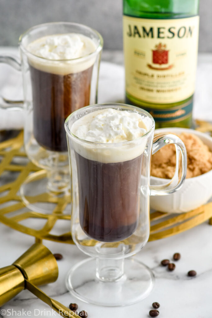 Overhead photo of two mugs of Irish Coffee topped with whipped cream. Bowl of brown sugar and bottle of Jameson Irish Whiskey on counter.