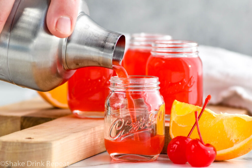 Close up photo of person's hand pouring Alabama Slammer Shots recipe into shot glasses. Orange slice and cherries sit beside shot glass.