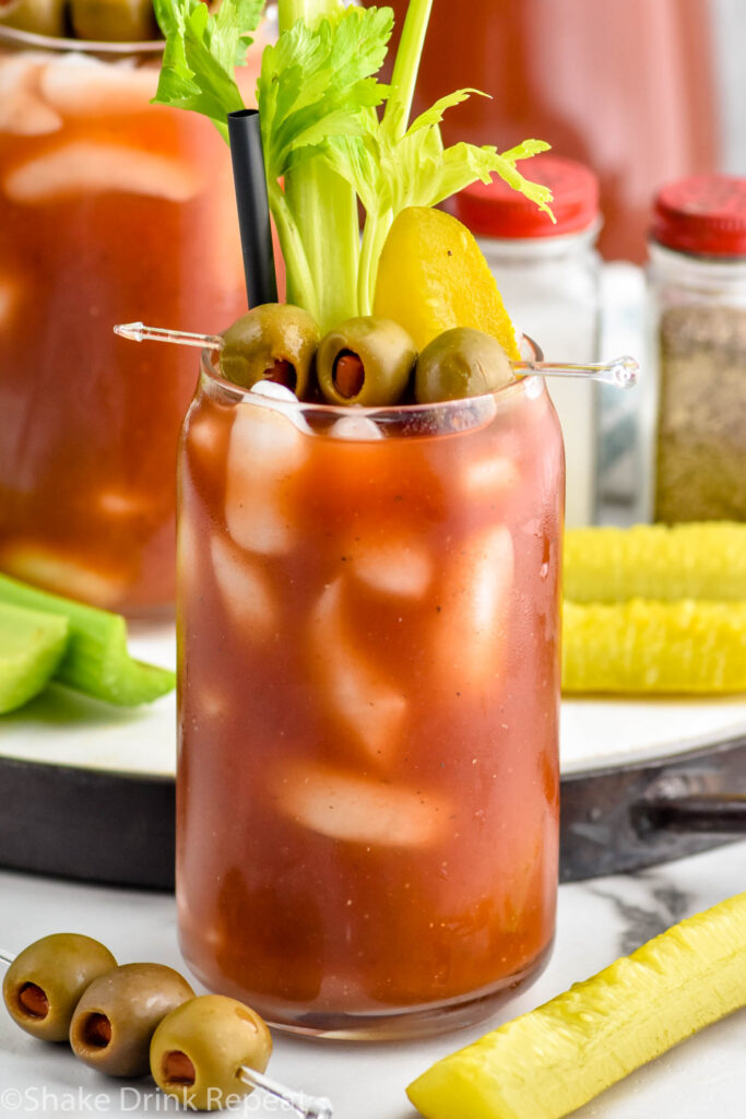 Overhead photo of Bloody Mary made Bloody Mary Mix and garnished with olives, celery, and a pickle. Extra olives, pickles, and celery beside glass.