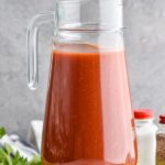 Photo of a pitcher of Bloody Mary Mix. Olives, celery, and a pickle on counter beside pitcher.