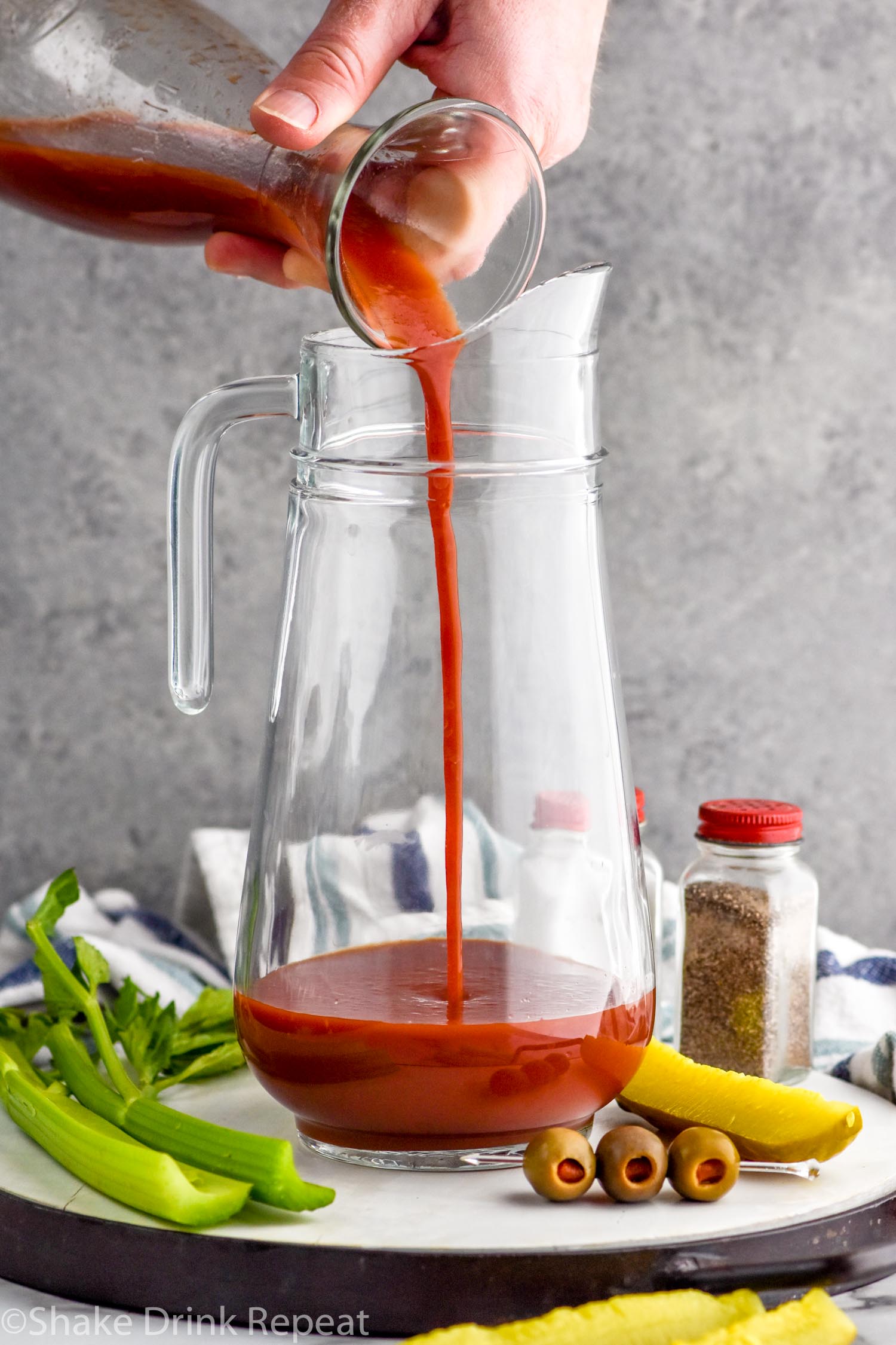 Photo of person's hand pouring tomato juice into pitcher for Bloody Mary Mix recipe. Olives, celery, and pickles on counter for garnish.