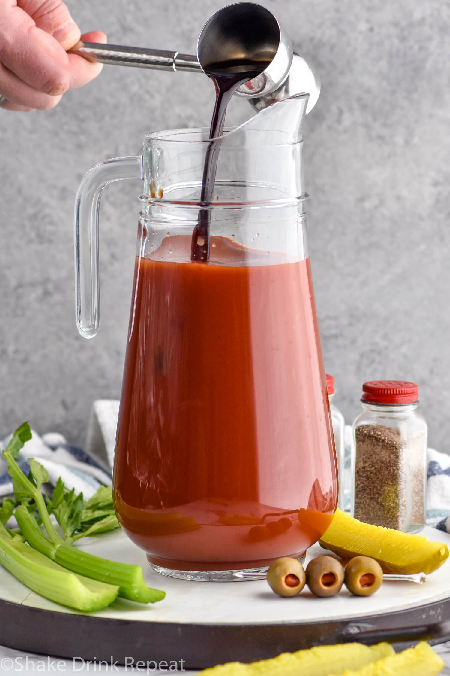 Photo of person's hand pouring worcestershire sauce into pitcher of ingredients for Bloody Mary Mix recipe.