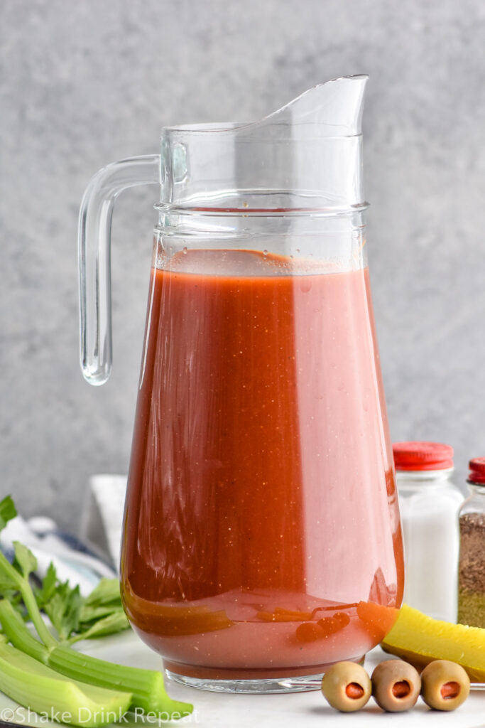 Photo of a pitcher of Bloody Mary Mix. Olives, celery, and a pickle on counter beside pitcher.