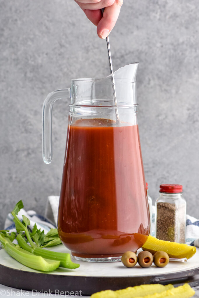 Photo of person's hand stirring pitcher of ingredients for Bloody Mary Mix recipe. Celery, olives, and pickle on counter beside pitcher for garnish.