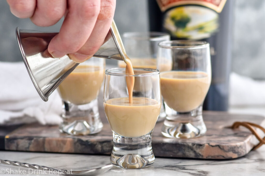 Photo of person's hand pouring Baileys Irish Cream into shot glasses for Cement Mixer Shot recipe. Bottle of Baileys is in the background.