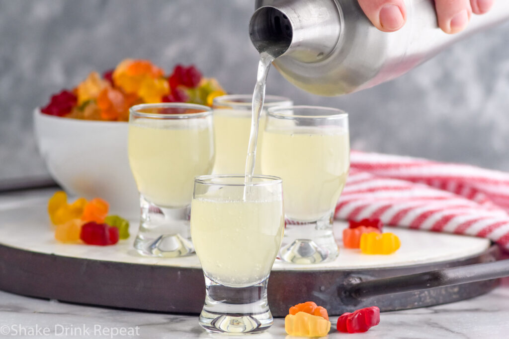 Photo of person's hand pouring Gummy Bear Shot recipe into shot glasses. Gummy bears on counter for garnish.