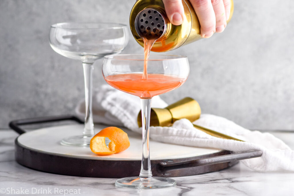 Close up photo of person's hand pouring a cocktail shaker of Paper Plane cocktail into coupe glass.