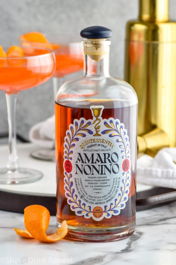 Close up photo of a bottle of Amaro Nonino. Two Paper Plane cocktails and a cocktail shaker in the background.