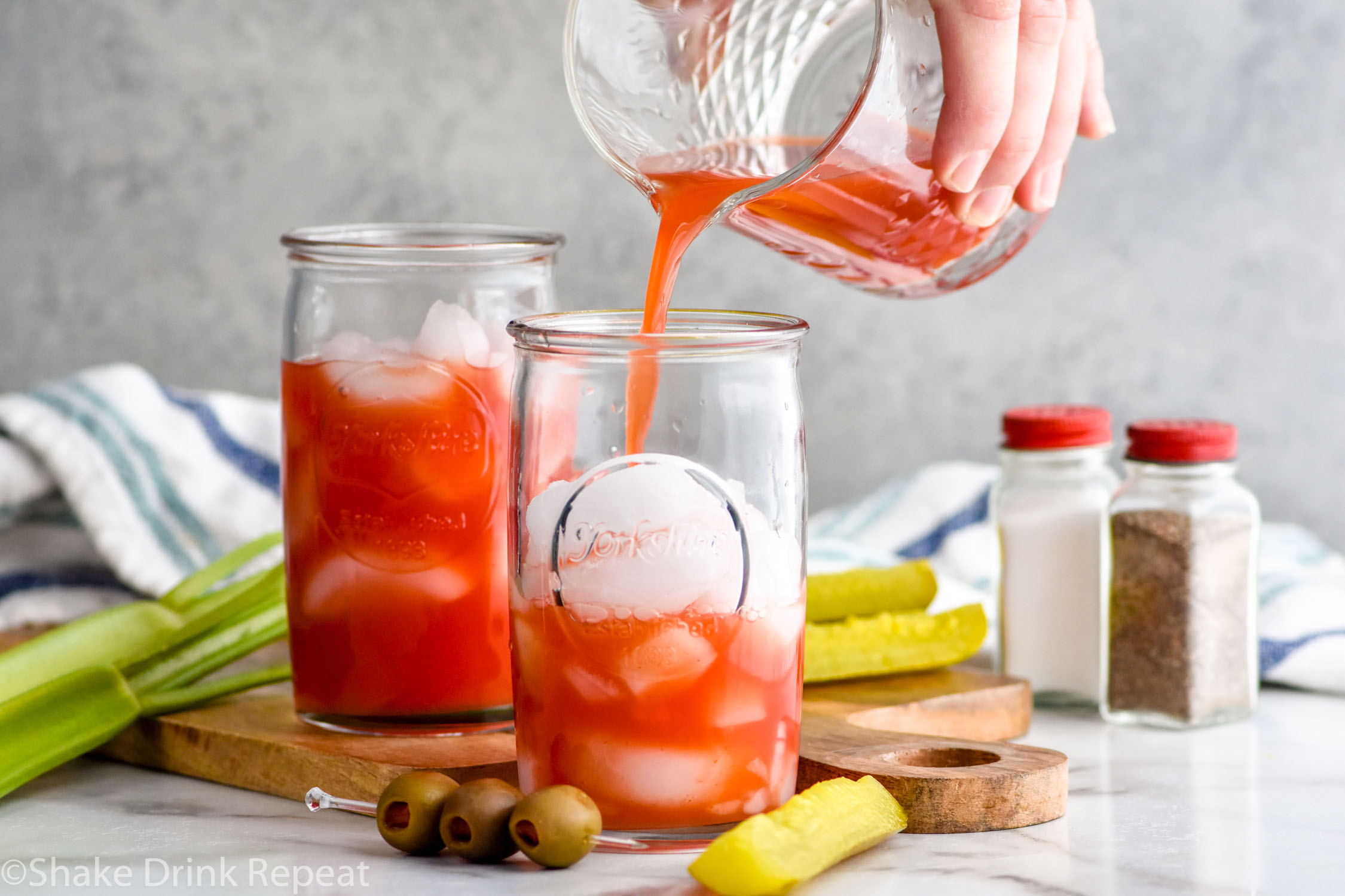 Photo of person's hand pouring Bloody Caesar recipe into glass of ice. Pickles, celery, and olives on counter beside glasses of Bloody Caesars. Salt and pepper shakers in the background.