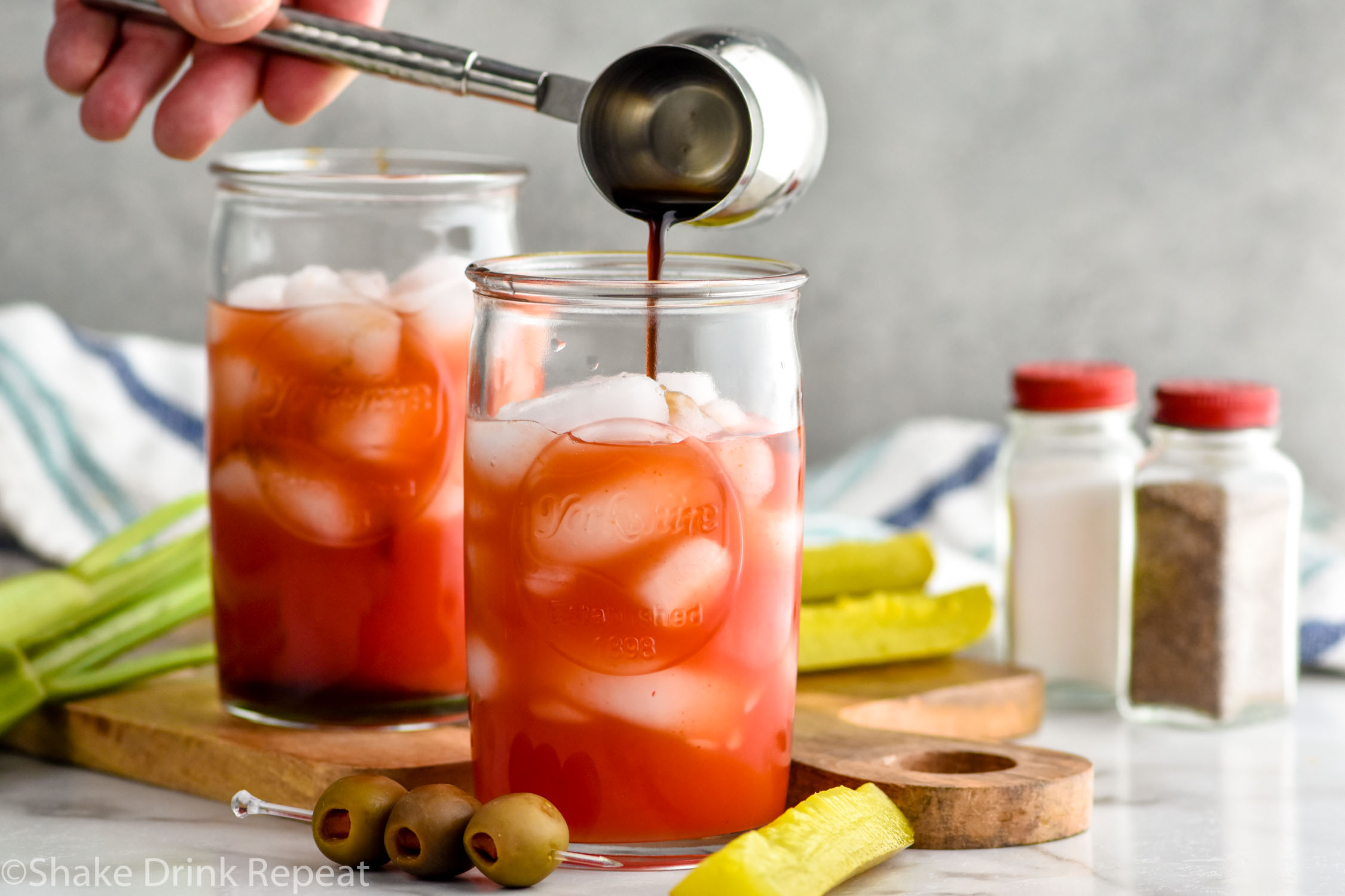 Photo of person's hand pouring Worcestershire sauce into glass of ice and ingredients for Bloody Caesar recipe. Celery, pickles, olives, salt, and pepper on counter for garnish. Another Bloody Caesar cocktail in background.