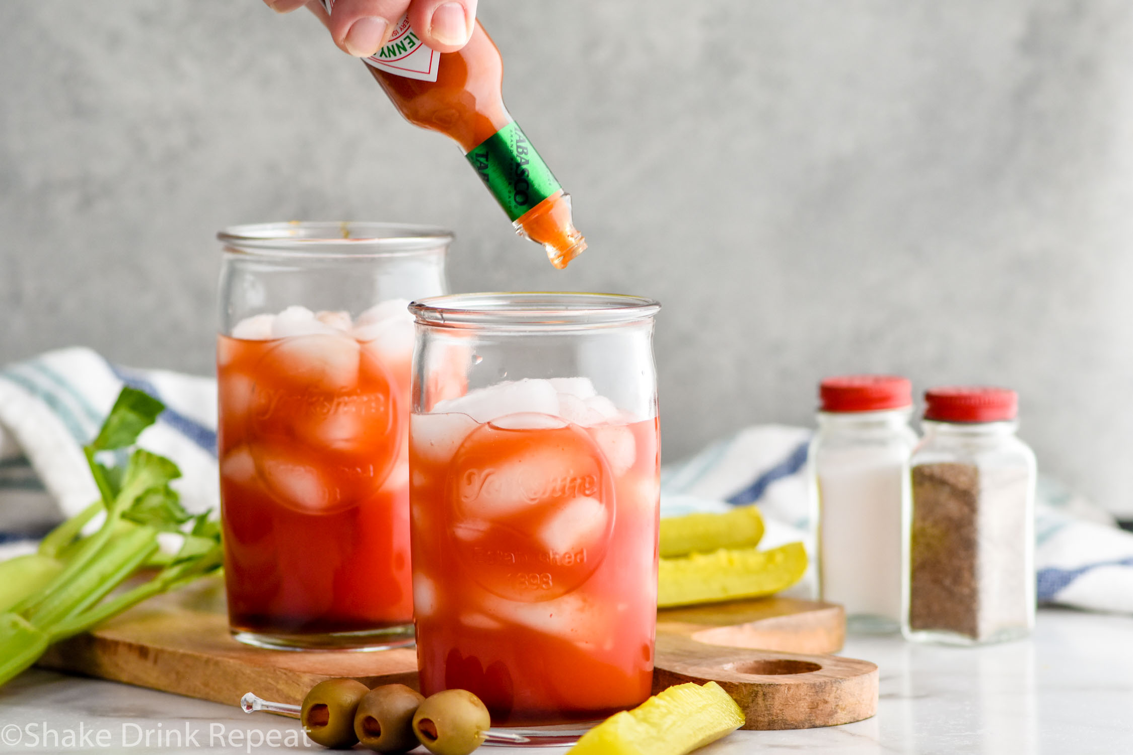 Photo of person's hand putting Tabasco sauce into glass of ice and ingredients for Bloody Caesar recipe. Pickles, olives, celery, salt, and pepper on counter for garnish.