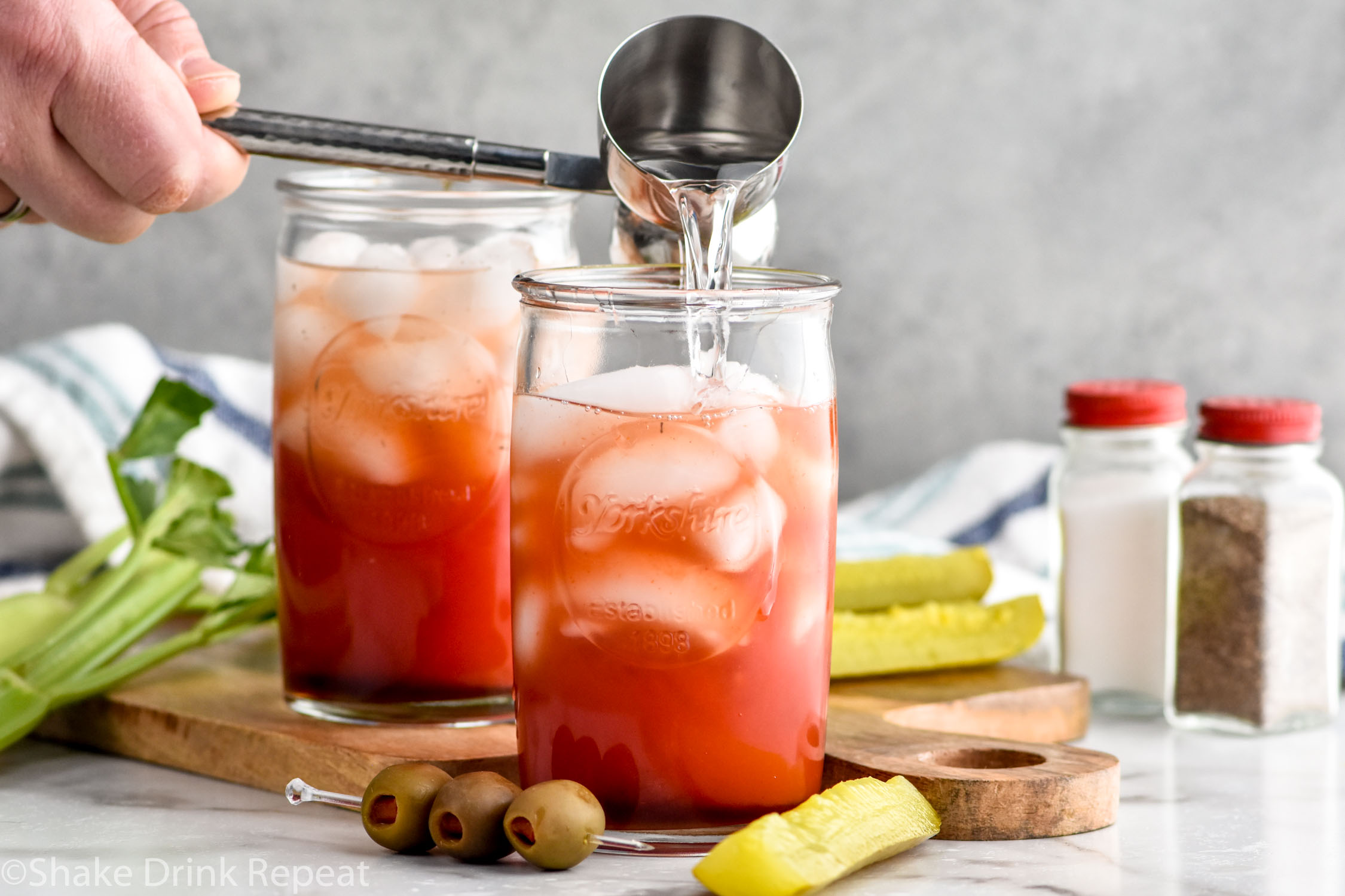 Photo of person's hand pouring vodka into glass of ice and ingredients for Bloody Caesar cocktails. Olives, pickles, celery, salt, and pepper on counter for garnish.
