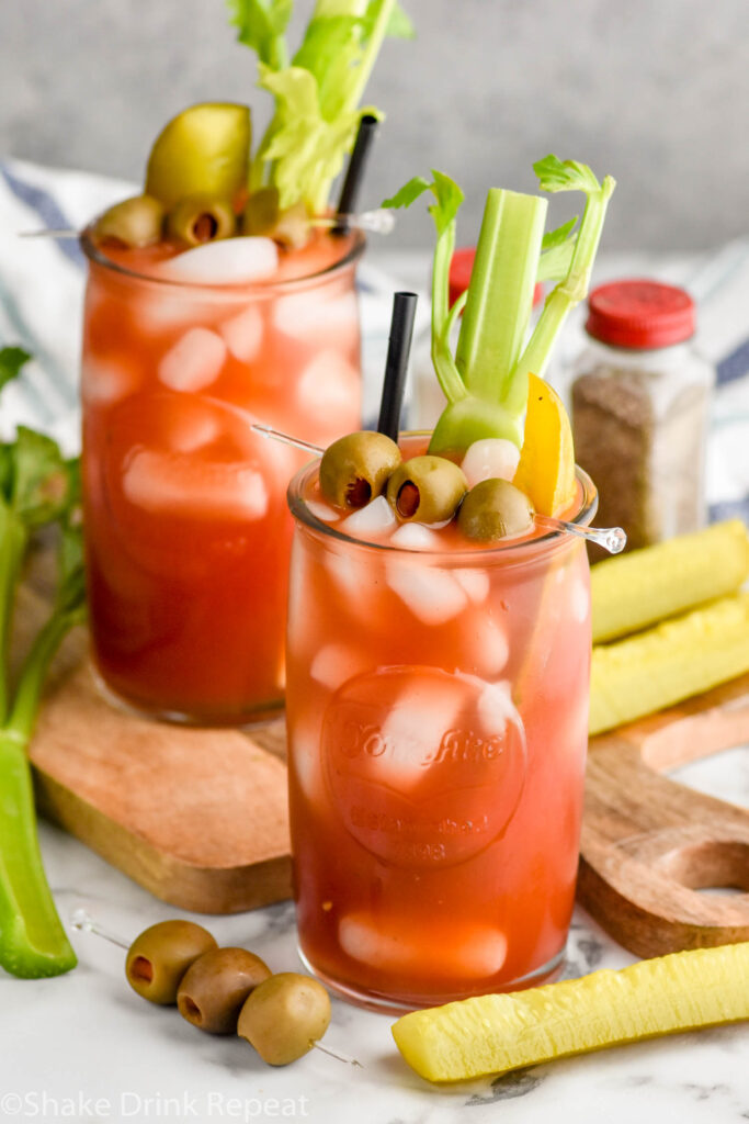 Overhead photo of Bloody Caesar cocktails garnished with olives, pickles, and celery, with extras for garnish on counter.