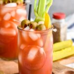 Photo of Bloody Caesar served in a glass garnished with pickles, olives, and celery. Extra olives and pickles on counter.