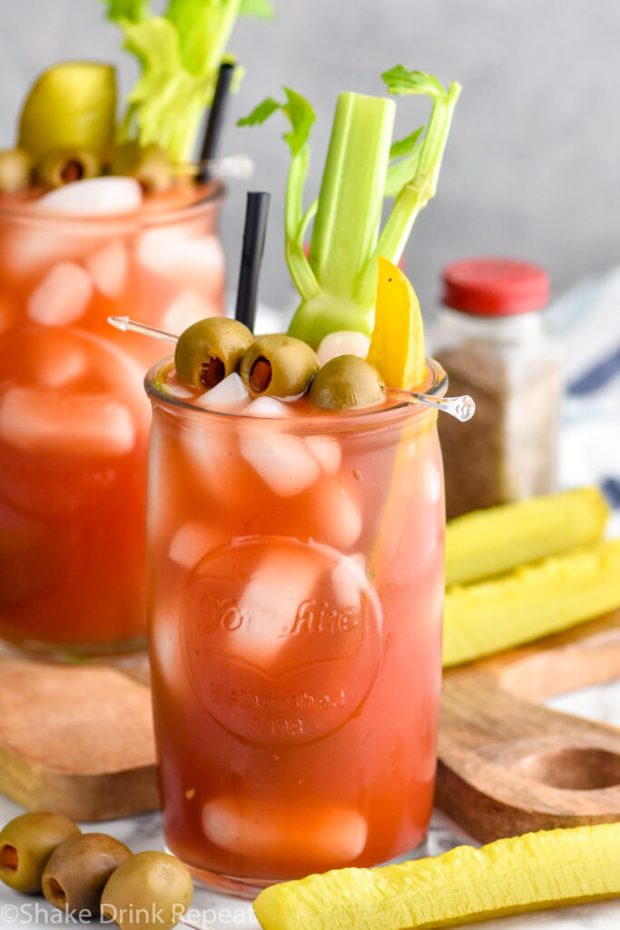 Photo of Bloody Caesar served in a glass garnished with pickles, olives, and celery. Extra olives and pickles on counter.
