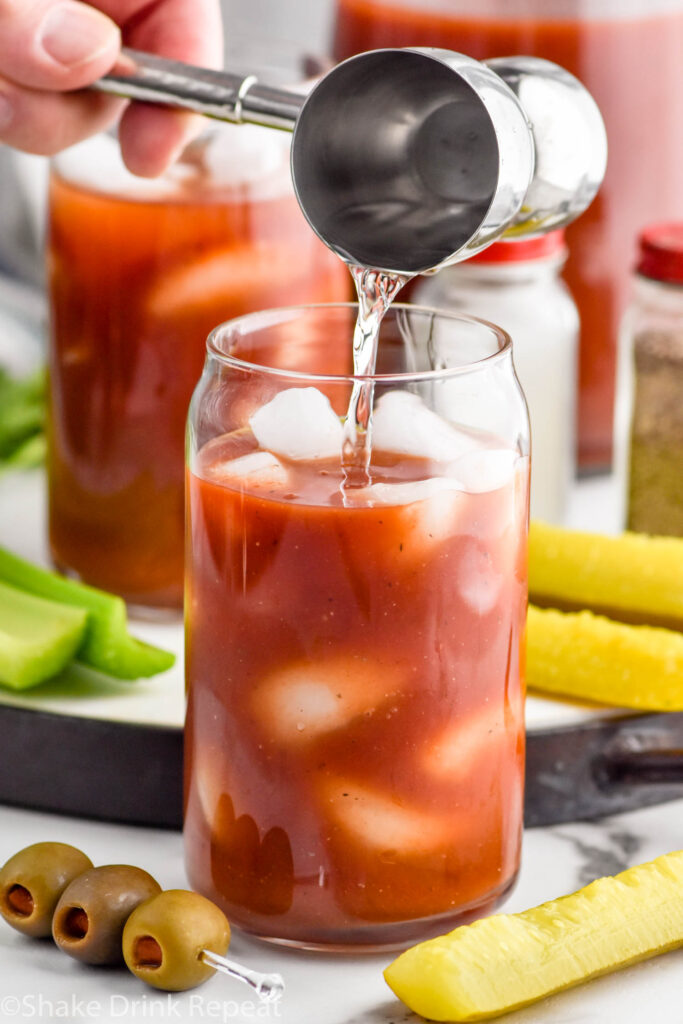 Close up photo of person's hand pouring vodka into glass of Bloody Mary mix for Bloody Mary recipe. Pickles, olives, and celery on counter beside glass for garnish.