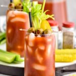 Photo of Bloody Marys garnished with olives, celery, and a pickle. Extra olives, celery, and pickles on counter beside Bloody Mary glass.