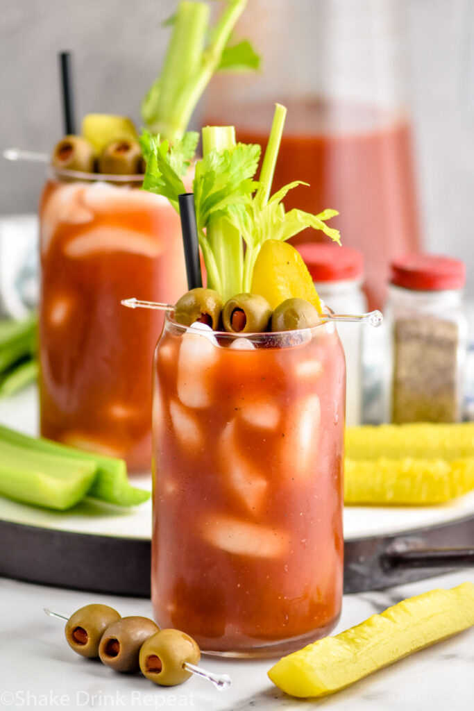 Photo of Bloody Marys garnished with olives, celery, and a pickle. Extra olives, celery, and pickles on counter beside Bloody Mary glass.