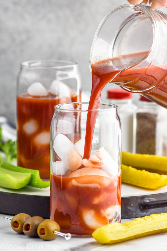 Photo of person's hand pouring pitcher of ingredients into glass of ice for Bloody Mary recipe. Pickles, olives, and celery on counter beside glass.