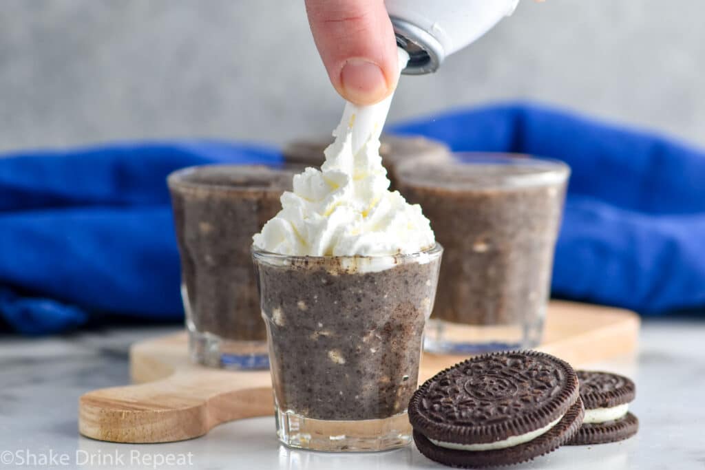 Close up photo of person's hand putting whipped cream on Cookies and Cream Pudding Shots. Oreo cookies on counter for garnish.