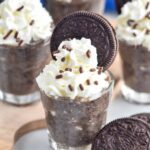 Photo of Cookies and Cream Pudding Shots garnished with whipped cream, sprinkles, and Oreo cookies. Extra Oreo cookies on counter for garnish.