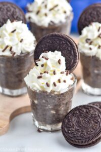 Photo of Cookies and Cream Pudding Shots garnished with whipped cream, sprinkles, and Oreo cookies. Extra Oreo cookies on counter for garnish.